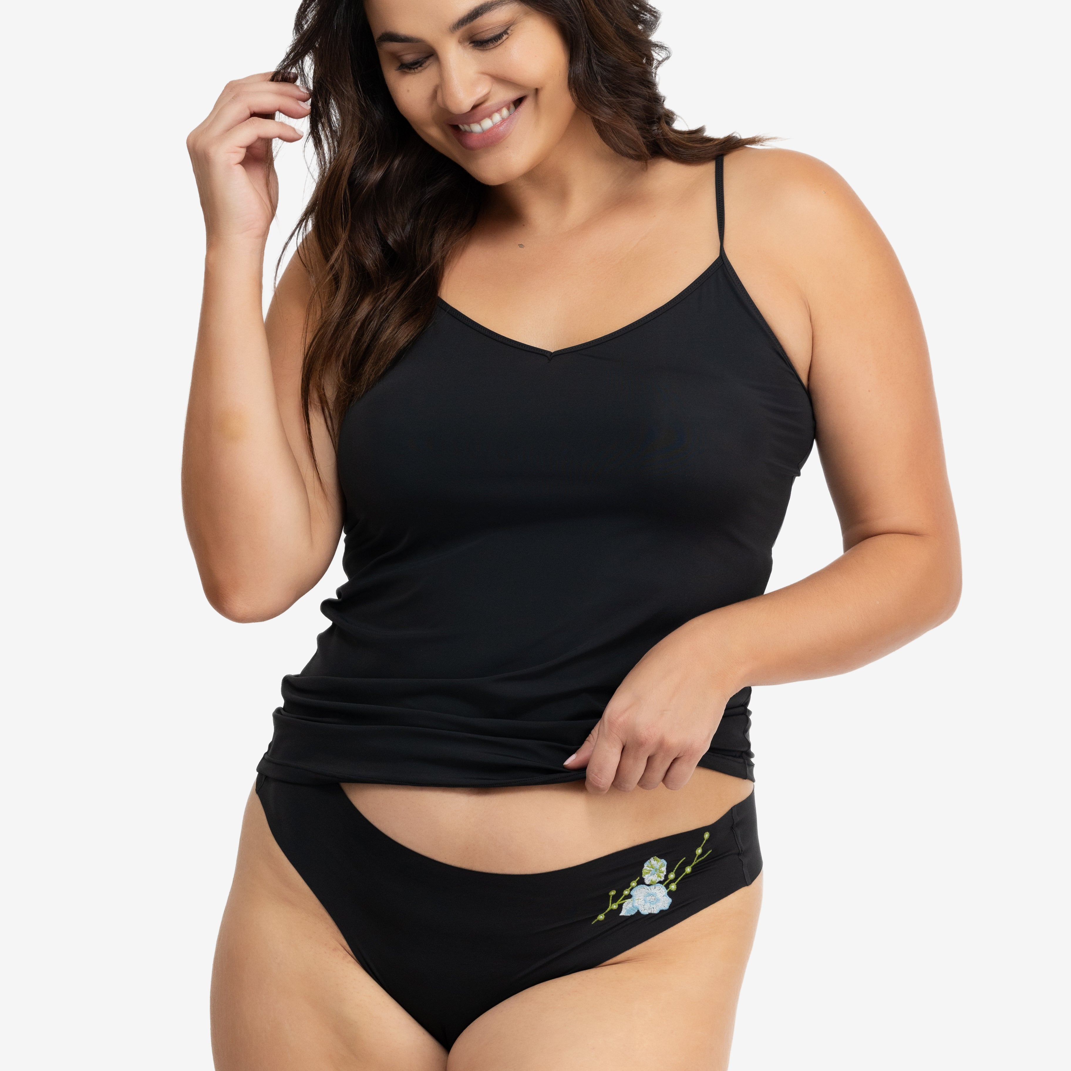 A woman wearing a soft seamless tap shoe black thong underwear with an embroidered floral pattern in contrasting colors against white background.