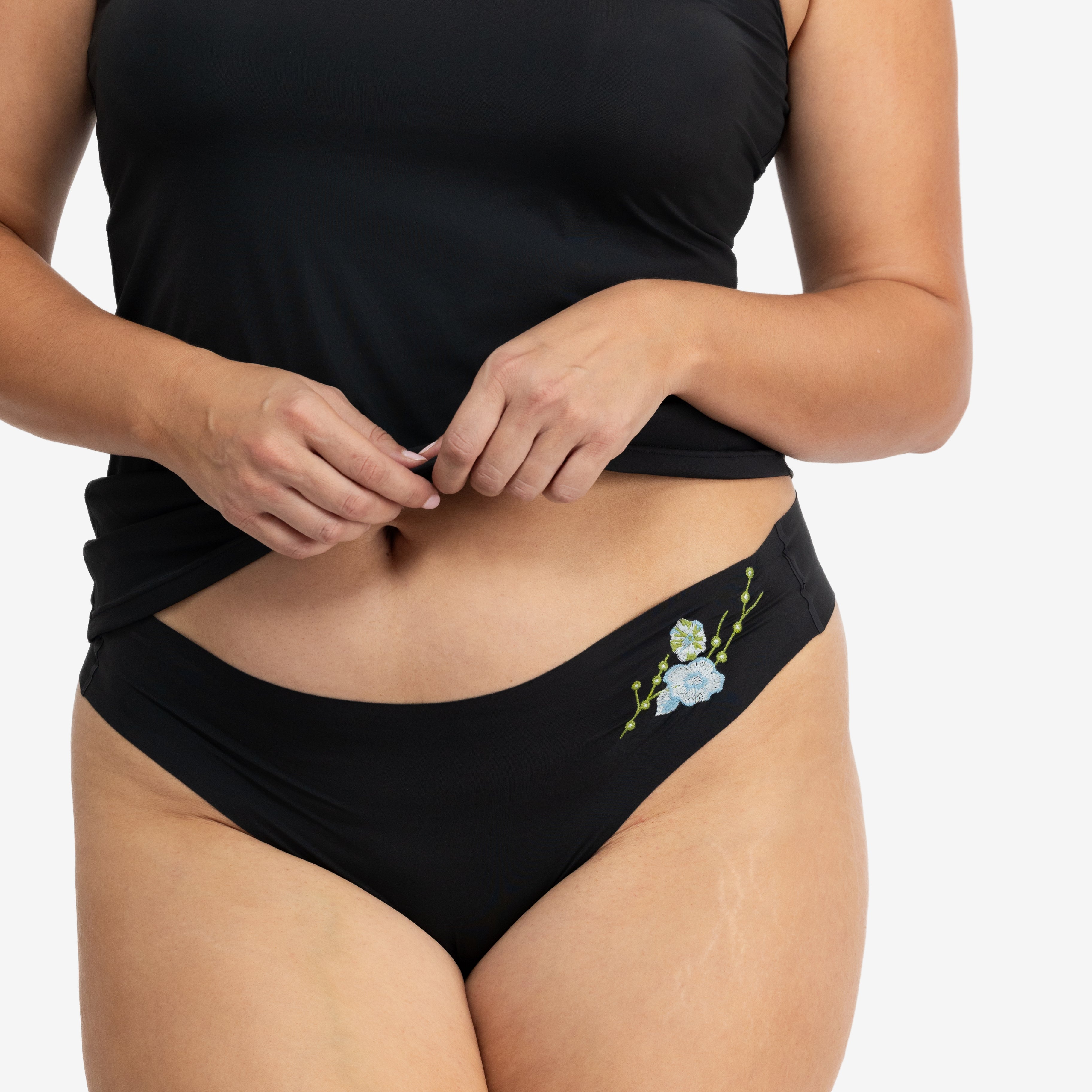 A woman wearing a soft seamless tap shoe black thong underwear with an embroidered floral pattern in contrasting colors against white background.