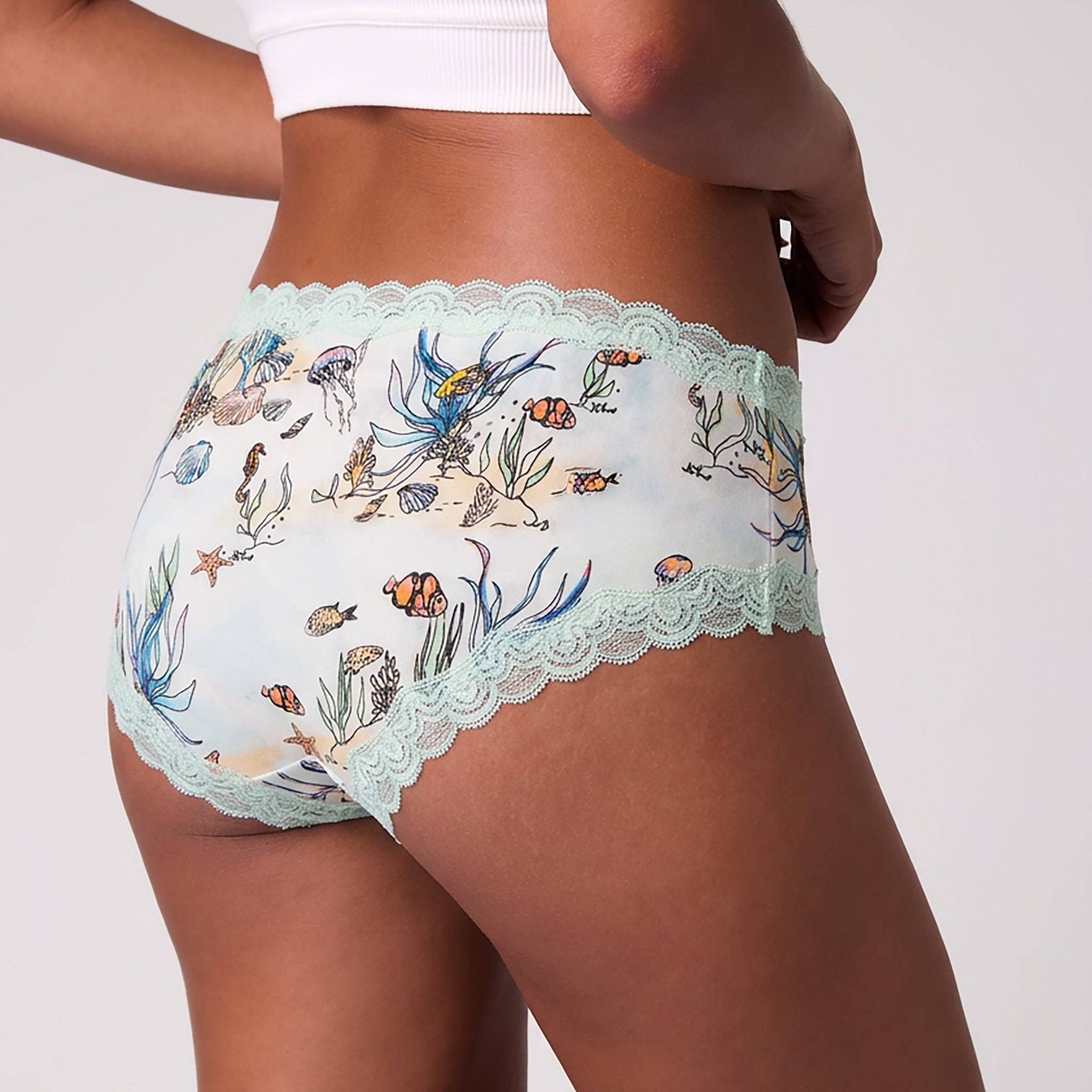 A model wearing Uwila Warrior Soft Silk Sea Print Briefs with light blue scalloped lace trim paired with a white ribbed crop top against a light gray background.