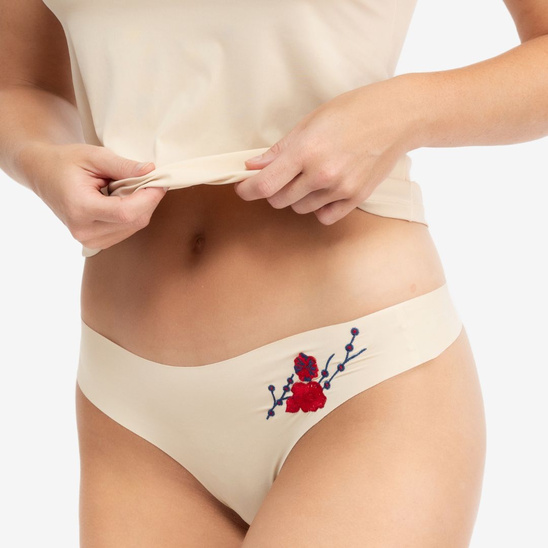 A woman wearing a soft seamless smoke grey thong underwear with an embroidered floral pattern in contrasting colors against white background.