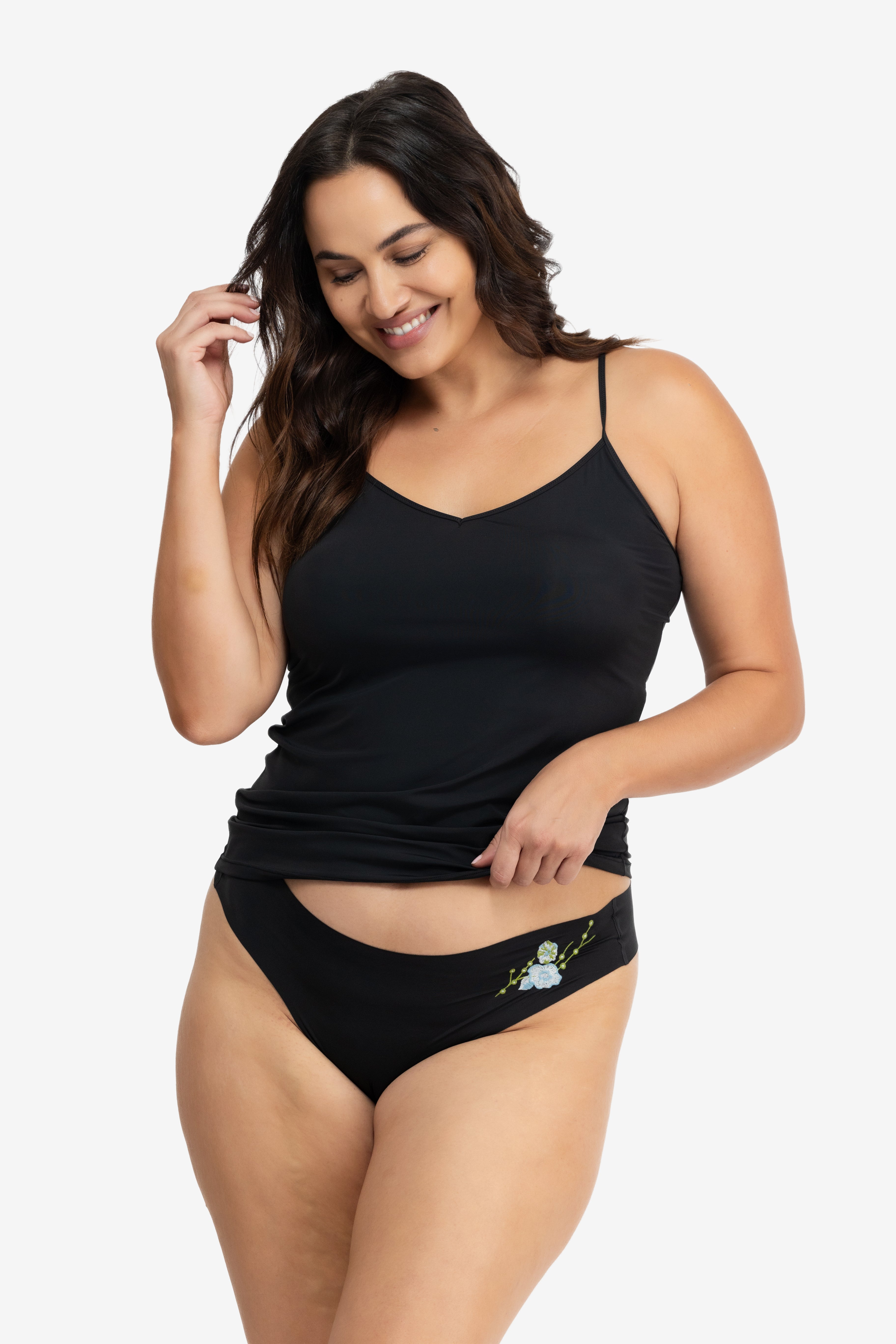 A woman wearing a soft seamless tap shoe black thong underwear with an embroidered floral pattern in contrasting colors against white background.