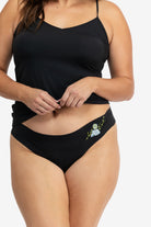A woman wearing a soft seamless tap shoe black thong underwear with an embroidered floral pattern in contrasting colors against white background.