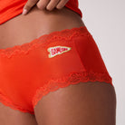 Close-up of a model wearing spicy orange lace-trimmed soft silk briefs with a small Game Day patch on the front