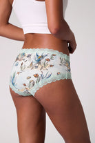 A model wearing Uwila Warrior Soft Silk Sea Print Briefs with light blue scalloped lace trim paired with a white ribbed crop top against a light gray background.