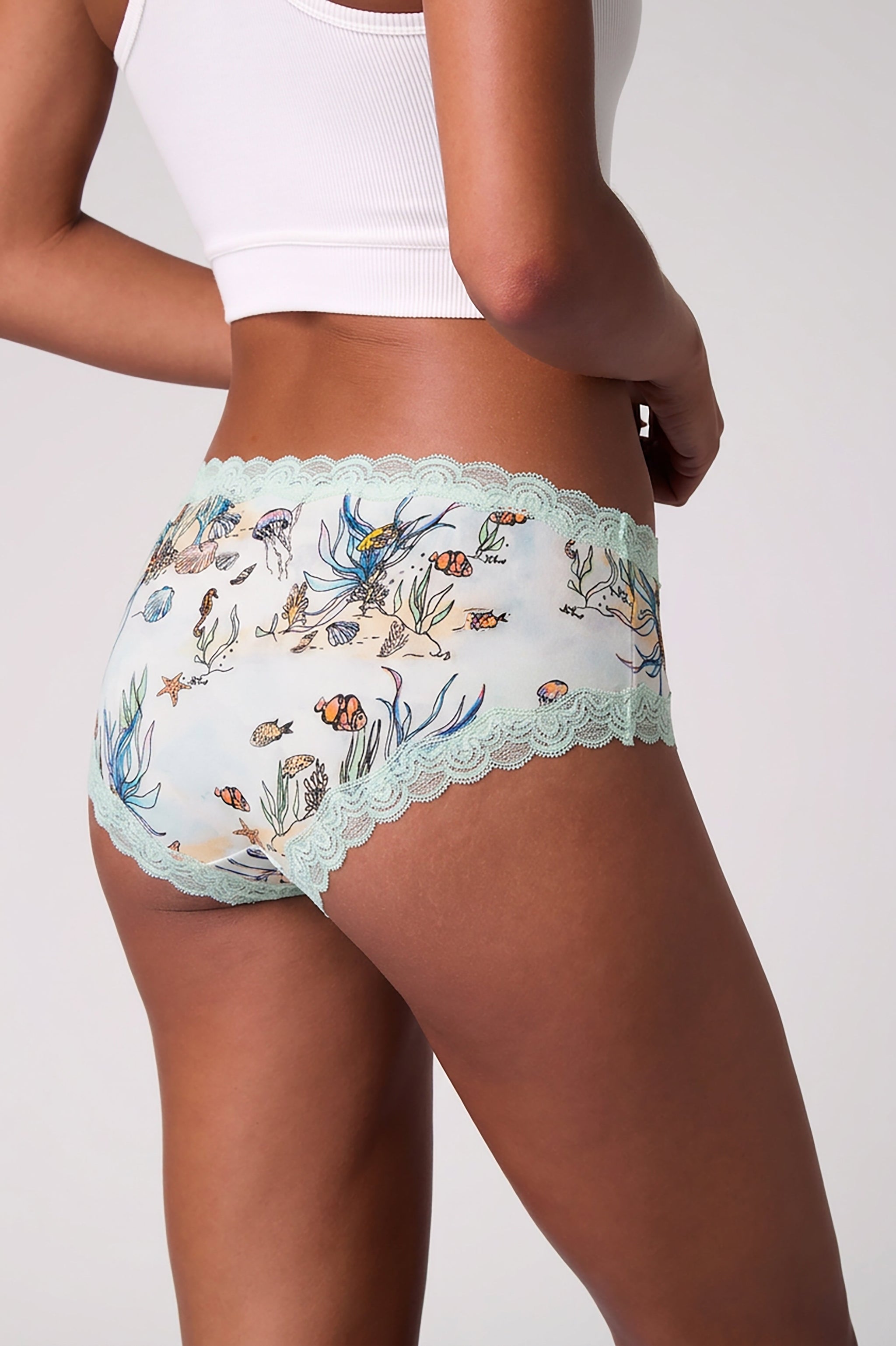 A model wearing Uwila Warrior Soft Silk Sea Print Briefs with light blue scalloped lace trim paired with a white ribbed crop top against a light gray background.