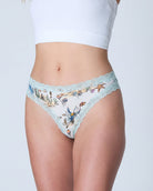 A model wearing Uwila Warrior Soft Silk Sea Print Thong with  lace-trimmed, paired with a white ribbed crop top against a light gray studio background.